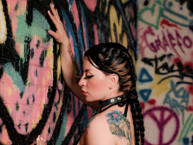 Katalina Johnson mit schwarzen, zu einem Zopf geflochtenen Haaren, trägt eine schwarze Halskrause, hat ein Tattoo an ihrer rechten Schulter und berührt mit ihrer linken Hand eine farbenfrohe Graffiti-Wand, im Seitenprofil, blickt nach unten..