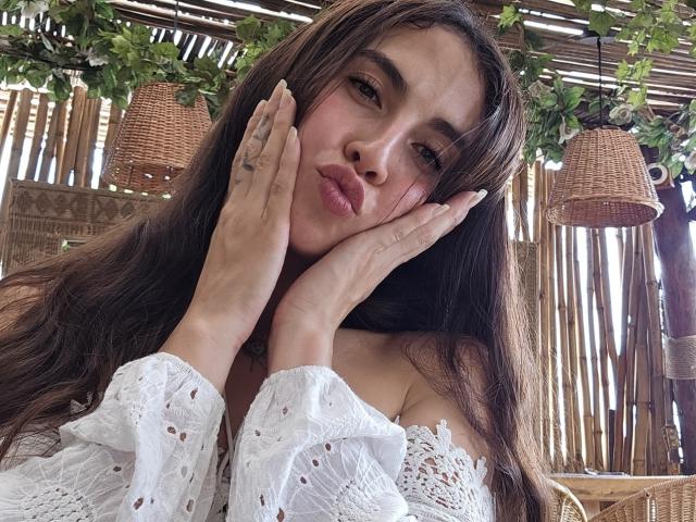 RossyNolen with long brown hair, white off-shoulder top, hands on face, pouty lips, brown eyes, tattoo on left hand, wicker hanging lamps above, green leaves in background.