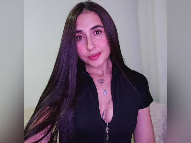 Rossy Nolen, con cabello largo y castaño oscuro, sombra de ojos rosa, una blusa negra con cremallera, pequeños tatuajes y una sonrisa sutil, mostrando ligeramente sus senos a través de la blusa..