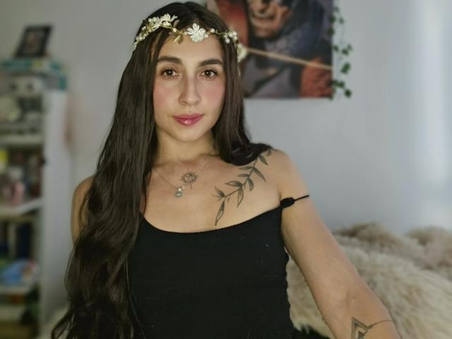 Rossy Nolen, con cabello castaño largo, usando una corona de flores, un top negro sin tirantes, mostrando tatuajes en el hombro y el pecho, sonriendo, labios rosados, ojos marrones, un collar pequeño, iluminación suave..