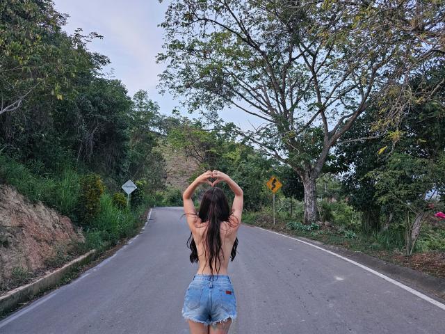RossyNolen, topless, with long brown hair, arms raised, wearing frayed blue denim shorts, standing on a road, back to camera, trees on both sides, dusk light.