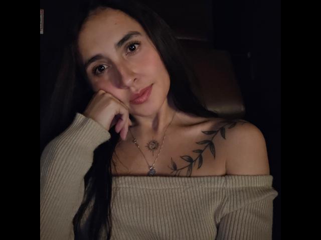 Rossy Nolen, con cabello negro largo, blusa beige de corte asimétrico, pechos pequeños, pecho tatuado, mano en la mejilla, sonrisa sutil, fondo oscuro, iluminación suave, collar con un dije de sol..