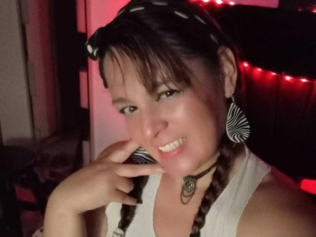 AmandaHotL with braided brown hair, white sleeveless top, black choker, zebra-pattern earrings, and a hand near her smiling face, red-lit background, and striped headband.