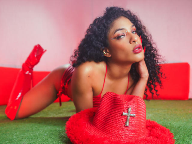 VioletHotX with curly black hair, red lipstick, and red boots, lying on green turf, wearing red lingerie, and a red hat with a cross, looking seductive.