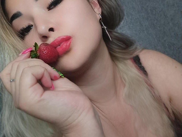 PussyXKitty with blonde hair, green nails, and cross earrings, sucking on a strawberry, lips pink and shiny, eyelashes long, wearing a silver ring on her right hand.