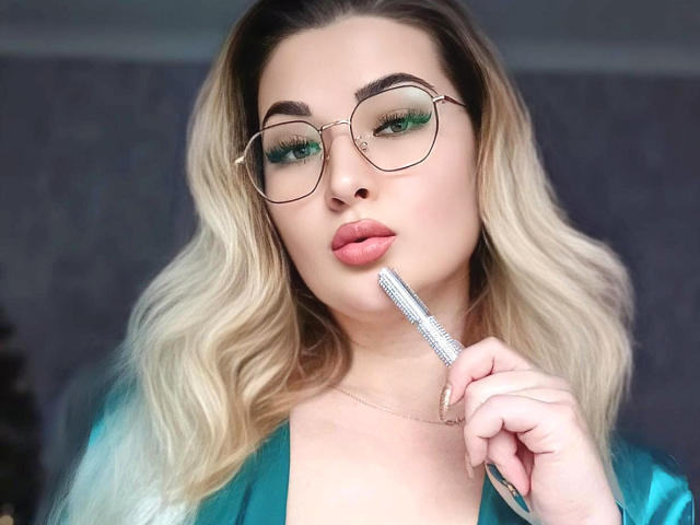 PussyXKitty with blonde hair, green eye makeup, and glasses, holding a brush to her pink lips, wearing a green satin top.