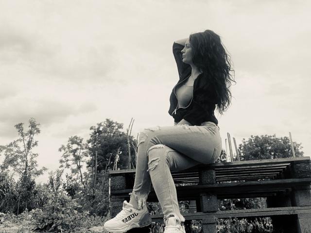 Coryna with long, wavy dark hair, unzipped black top showing her tits, ripped jeans, and white Adidas sneakers, sitting on a wooden bench, hand in hair, looking side.