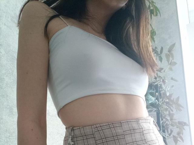WollyMolly with small tits in a white, one-shoulder crop top and beige plaid pants, showing her flat stomach and part of her brown hair.