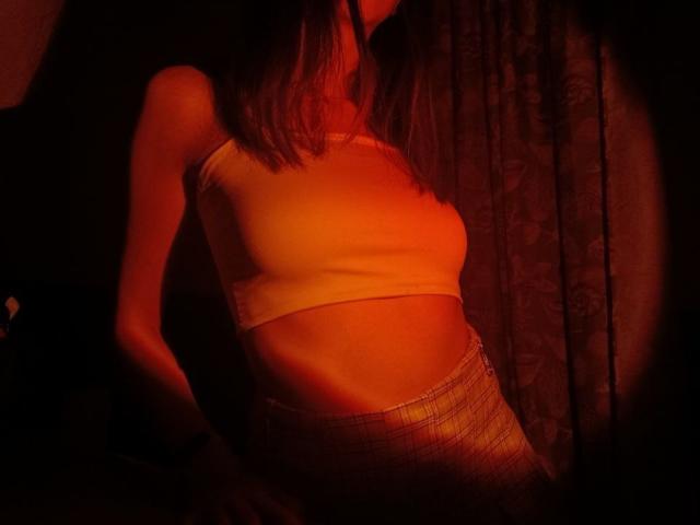WollyMolly with red-orange lighting, wearing a white sports bra, showing small tits and a slim, toned belly, in a dark, textured background. WollyMolly has long brown hair.