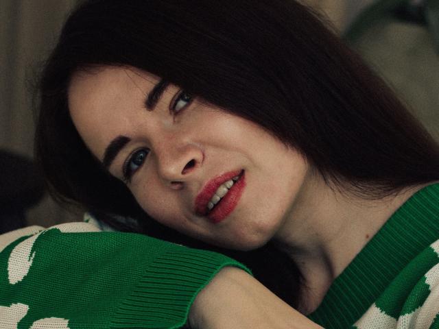 JasmineKouch, com cabelo castanho escuro, olhos verdes e batom vermelho, apoiada em um suéter verde e branco, sorrindo levemente. Seu rosto está inclinado para a direita..