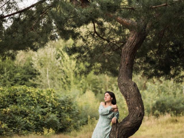 JasmineKouch apoyada contra un árbol retorcido, vestida con un vestido azul de un solo hombro, con ojos verdes y cabello oscuro, en un bosque soleado. Sus senos son ligeramente visibles..