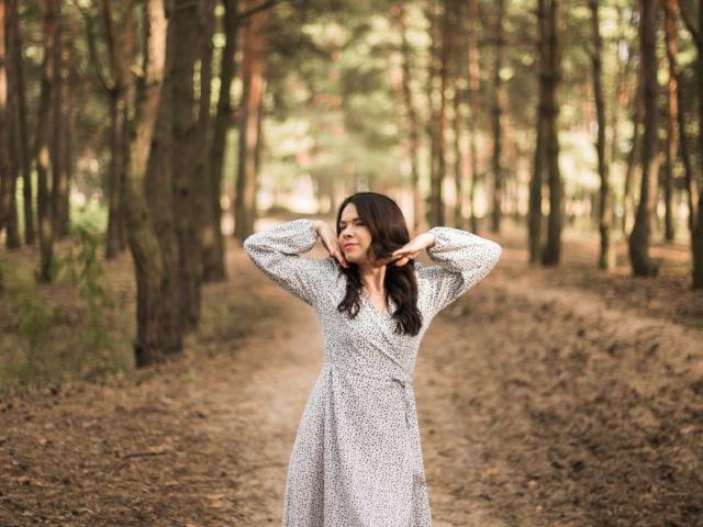JasmineKouch está de pie en un bosque, vestida con un vestido blanco de manga larga y estampado floral, con las manos en su cabello ondulado y castaño oscuro, sonriendo suavemente..