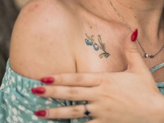 JasmineKouch's tits with a small blue and green flower tattoo on her right shoulder. Her red-nail-painted hand touches her chest, wearing a teal, patterned dress.