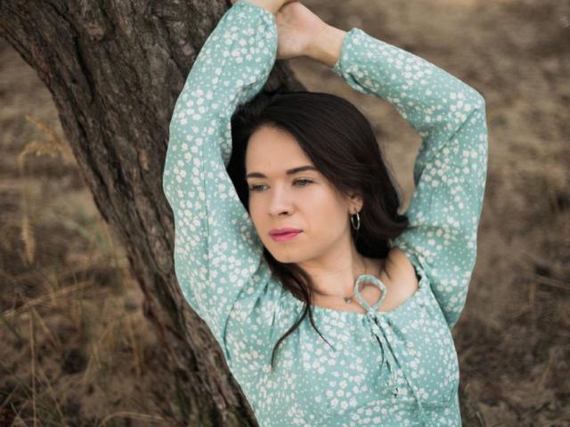 JasmineKouch with arms above head, leaning against tree, wearing teal floral dress, small hoop earrings, pink lipstick, dark hair, soft expression, standing in grassy, dry area.