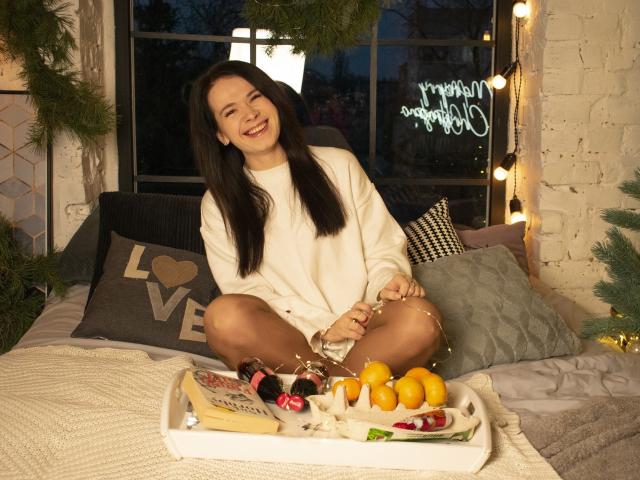 JasmineKouch with dark hair, white sweater, sitting cross-legged on bed, smiling, holding wine glass, Christmas tray with cheese, grapes, oranges, and candles in front of her.