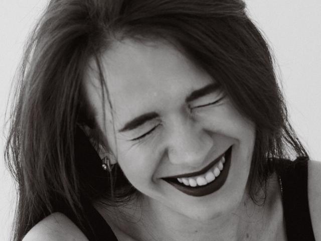 JasmineKouch tiene un lápiz labial oscuro, los ojos cerrados y una gran sonrisa que muestra los dientes. Lleva una camiseta de tirantes negra, y su cabello largo y despeinado cubre ligeramente su rostro..