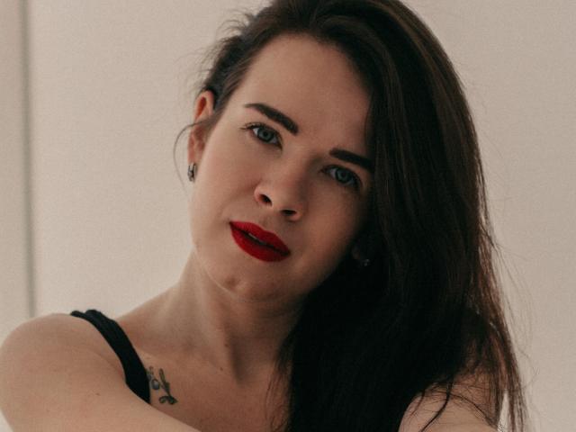 JasmineKouch a de longs cheveux bruns, du rouge à lèvres, des boucles d'oreilles noires et un tatouage sur son épaule droite. Elle porte un haut noir et regarde l'appareil photo avec un léger sourire..