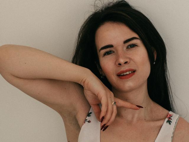 JasmineKouch, con i capelli scuri, il rossetto rosso e l'abito bianco con fiori, alza un braccio, mostrando l'ascella, con le unghie rosse, un anello e un sorriso. È carina, con un trucco naturale e un leggero sorriso..