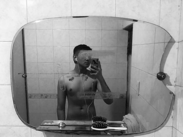 Black-and-white mirror selfie of Dekk, shirtless, with headphones in, tattoos on chest, holding phone, small boobs, flat ass, short dark hair, comb on mirror, tiled bathroom.
