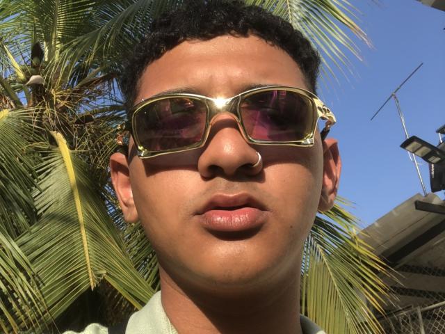 Dekk with curly black hair, tan skin, nose ring, and gold sunglasses, has pink-tinted lenses, glossy pink lips, and a green shirt, standing in front of a palm tree.