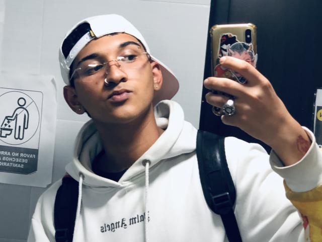Dekk with glasses, nose ring, and white cap, taking a selfie in a mirror. Wearing a white hoodie, backpack, and earbuds. Holding a phone with a colorful case.