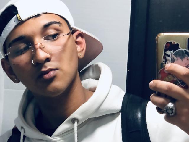 Dekk with glasses, white hoodie, and pink baseball cap, holding phone with ring, backpack, and gold case, has nose ring, earrings, and a slightly pouty expression.
