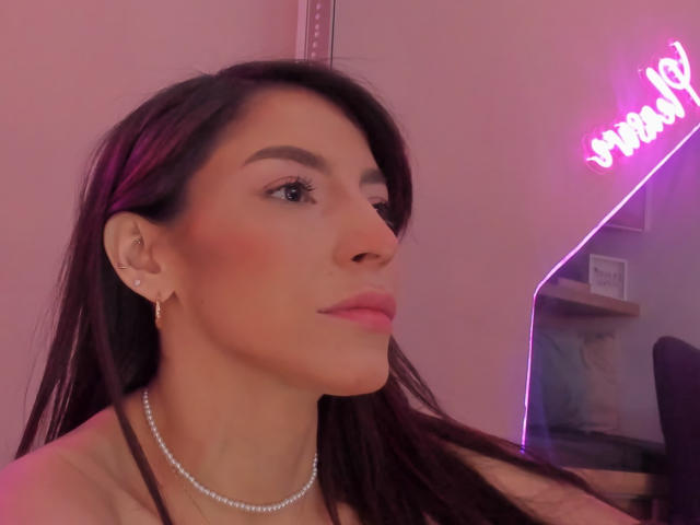 ValenRosee has dark brown hair, pink lipstick, and a pearl necklace, with small earrings, and a confident, slightly pouty expression, looking to the right. Neon pink light in background.