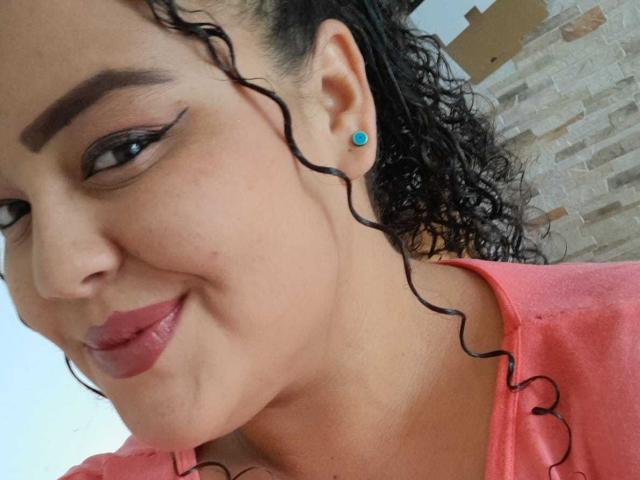 MelannieCarter has curly black hair, dark eyebrows, blue earbuds, and pink lipstick. She's wearing a pink shirt, smiling with her ass barely visible in the background.