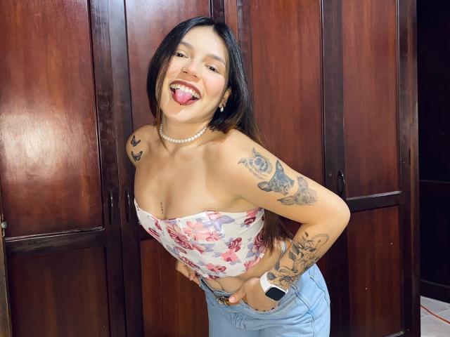 SophiaMarx, com longos cabelos pretos, língua de fora, vestindo um top floral sem alças, jeans azuis, um colar de pérolas, tatuagens nos braços e um smartwatch, está em frente a armários de madeira..