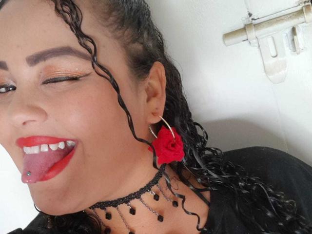 MelannieCarters Gesicht, mit einem Auge zwinkernd, trägt roten Lippenstift, eine schwarze Halskette und rote Ohrringe. Sie steckt ihre Zunge heraus und zeigt ein Zungenspiering. Ihr Haar ist lockig..