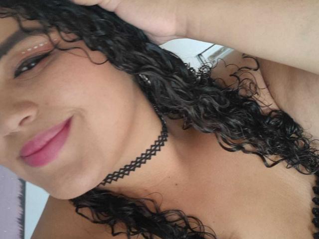 MelannieCarter with curly black hair, pink lipstick, and black choker, smiling, hand in hair, showing bare shoulders, tan skin, and subtle makeup. Boobs partially visible in frame.