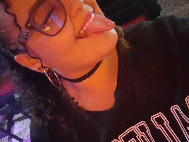 Le visage de Melannie Carter, la langue tirée par un piercing, portant des lunettes, un collier noir et des boucles d'oreilles cerceaux, est visible sur une photo où elle porte une chemise noire. Ses cheveux bouclés et son expression espiègle sont également visibles..