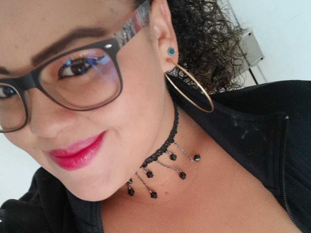 MelannieCarter with curly brown hair, black-framed glasses, red lipstick, gold hoop earrings, black necklace with black beads, black jacket, and blue ear piercing, smiling at the camera.