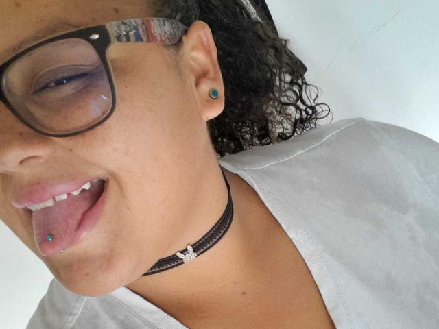 MelannieCarter's face, showing her brown skin, curly black hair, black glasses, blue ear studs, black choker with a silver charm, and her tongue with a blue piercing.