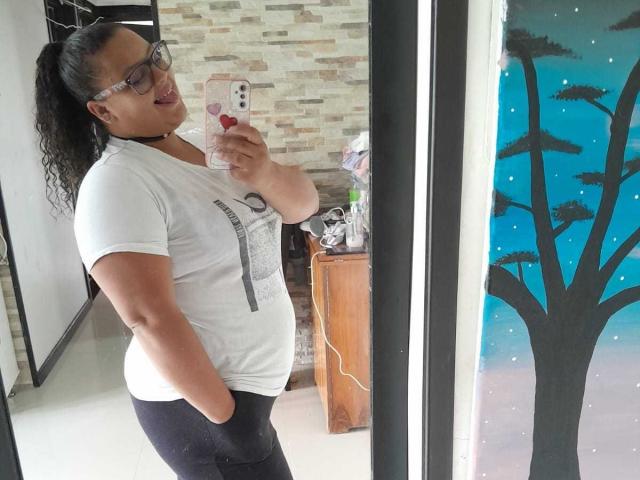 MelannieCarter with curly black hair, glasses, white tee, black pants, holding phone, tongue out, standing sideways, reflecting in mirror, showing her ass in the photo.