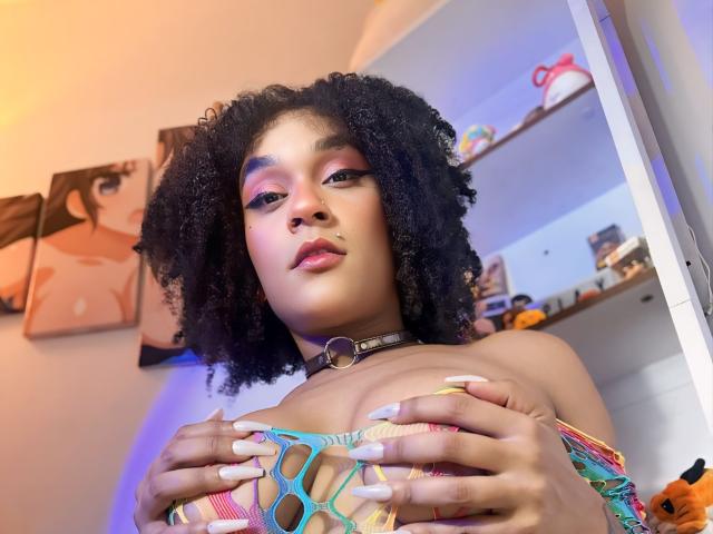 AmyStone69 with curly black hair, wearing a colorful mesh top, covering her tits with hands, choker, pink and blue eyeshadow, pink lips, and long white nails, in a bedroom.