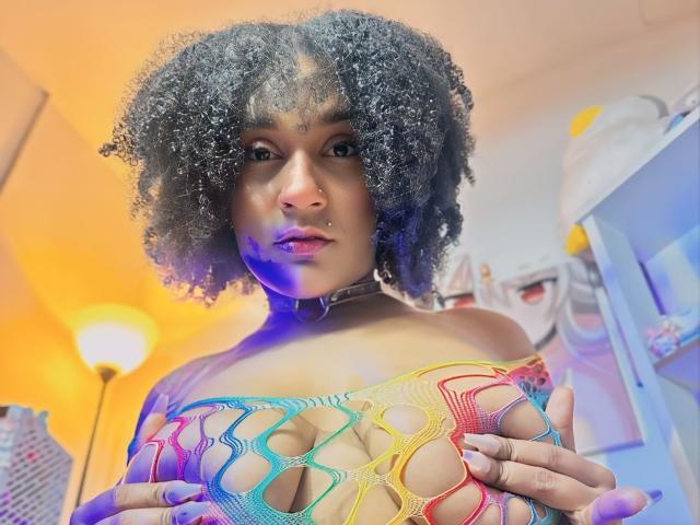 AmyStone69 with curly black hair, nose piercing, colorful netted top, holding her tits, purple and blue lighting, in a room with a standing lamp and wall art.
