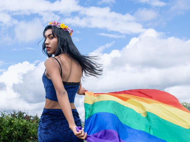 Kattaleya, cheveux foncés, couronne de fleurs, haut court en velours bleu et jupe, tenant un drapeau arc-en-ciel, dos à la caméra, ciel bleu, nuages duveteux, buissons verts, le vent agitant le drapeau..
