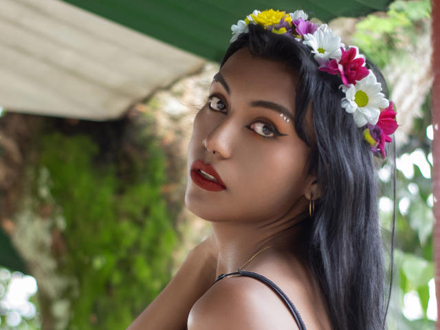KattaleyaHorny with a flower crown, dark hair, and red lipstick, looking back with arched eyebrows and gold earrings. Boobs visible, brown eyes, and a slight smile.