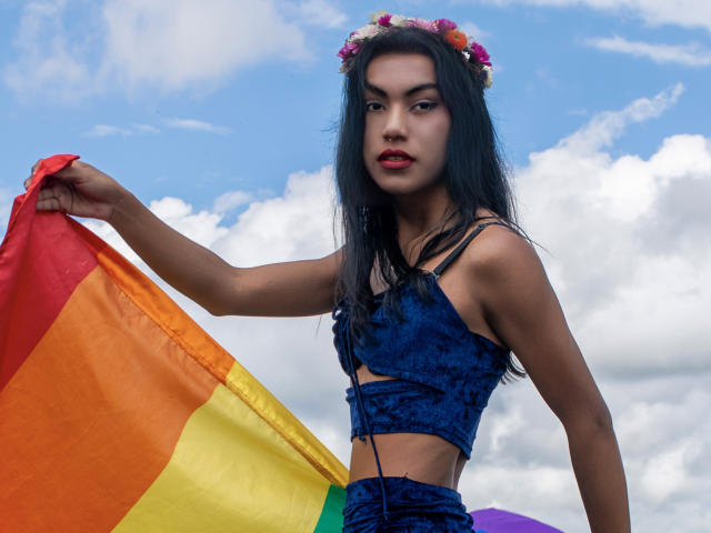 KattaleyaHorny with long black hair, wearing a blue velvet crop top, flower crown, and holding a rainbow flag, showing a confident, sexy, and attractive look.