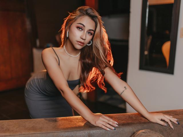 Alisses with long, wavy brown hair, wearing a tight grey dress, leaning on a brown surface, showing her small tits and ass, wearing a necklace, and a tattoo on her arm.