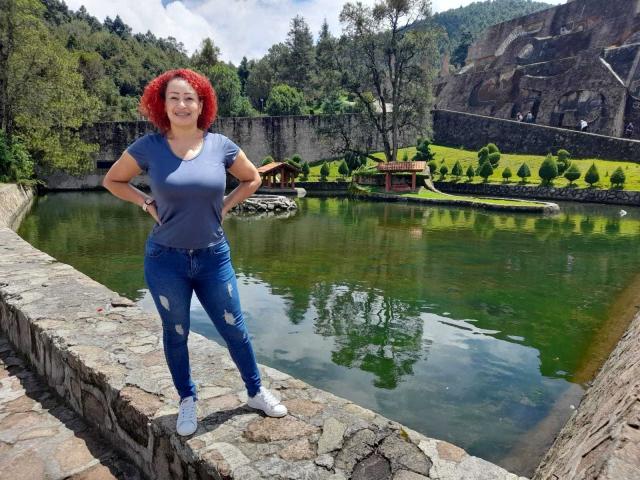 EliSheva stands with hands on hips, wearing a blue shirt, ripped jeans, and white sneakers, with red curly hair, by a green, reflective pond, on a stone wall.