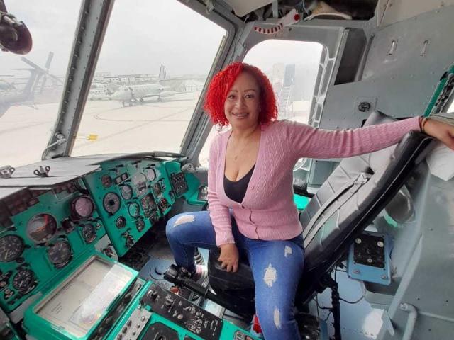 EliSheva with red curly hair, pink cardigan, black tank top, and ripped blue jeans, sits in a cockpit, smiling, right arm on seat, left hand on panel.