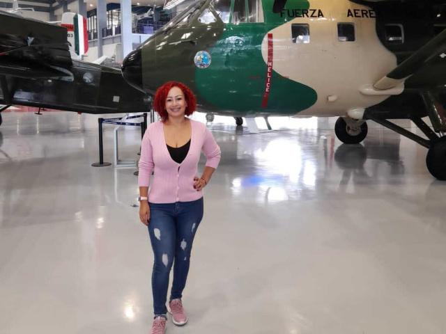 EliSheva with red hair, pink cardigan, black bra, ripped blue jeans, and pink sneakers, standing in front of a green military helicopter, smiling.