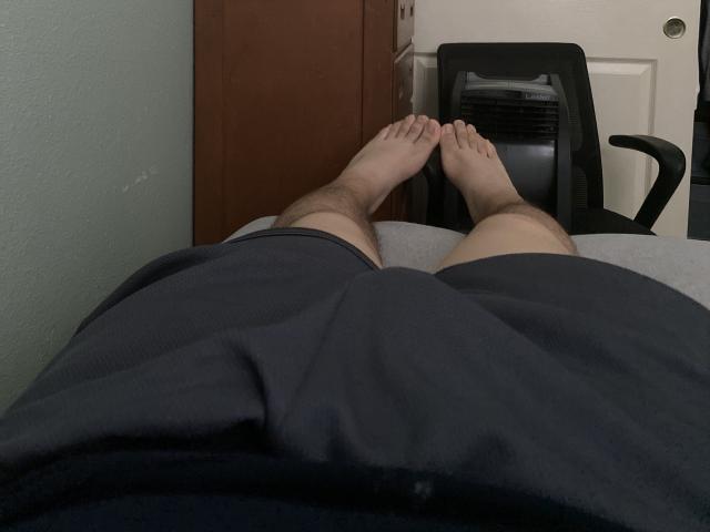 HornyDickx's legs, bare feet, crossed at the ankles, wearing dark blue shorts, lying on a bed, with a black office chair and door in the background.
