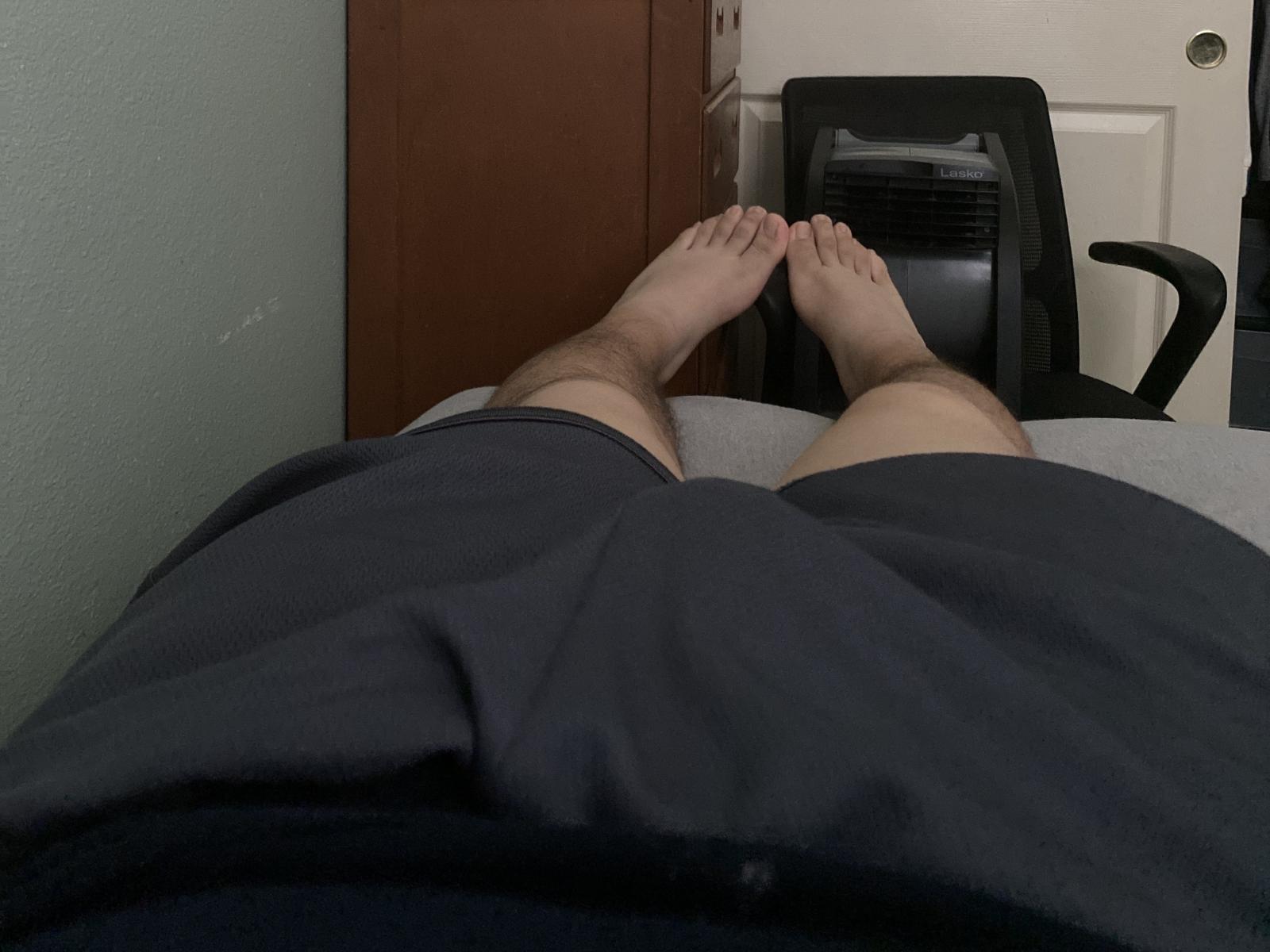 HornyDickx's legs, bare feet, crossed at the ankles, wearing dark blue shorts, lying on a bed, with a black office chair and door in the background.