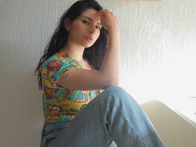 JeneKurz with long black hair, wearing a colorful cartoon-patterned top and blue jeans, sitting with one knee up, hand on head, looking at camera with a slight smile.