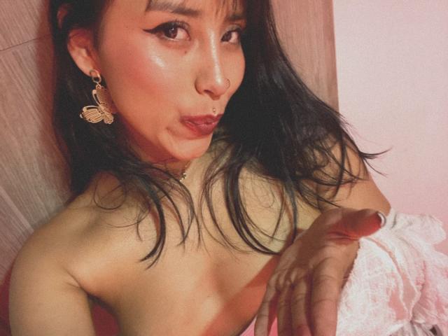 ElizabethMacy with black hair, pouty lips, and gold butterfly earrings, holding her bare tits, finger pointed, wearing a pink lace cloth, in a soft pink-lit room.