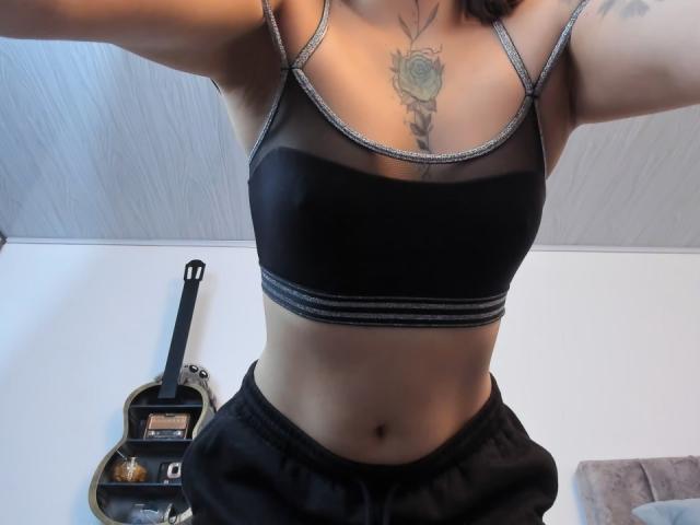 KayleeM with tan skin, black sports bra, and black pants, showing her small tits, flat stomach, and rose tattoo on her chest. Guitar and shelf in the background.