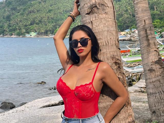 Maddison Aderson, com cabelo escuro, body de renda vermelho, seios grandes, óculos de sol pretos, em pé contra uma palmeira, com a mão na cabeça, com aparência sexy, perto da água, vestindo jeans azuis..